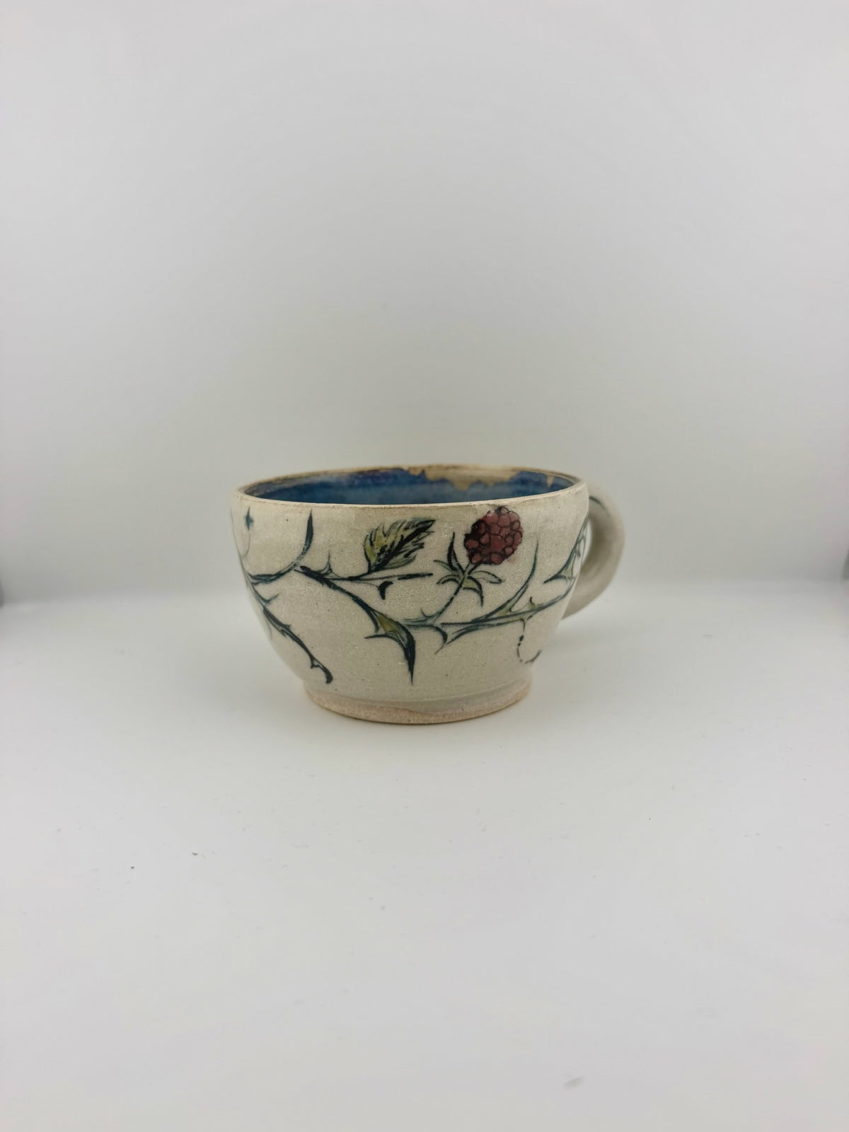 A handmade ceramic coffee cup with a cream-colored exterior, decorated with a hand-painted botanical design of green leaves and a red berry. The interior of the cup is glazed in a deep blue, and the handle is integrated into the side design.
