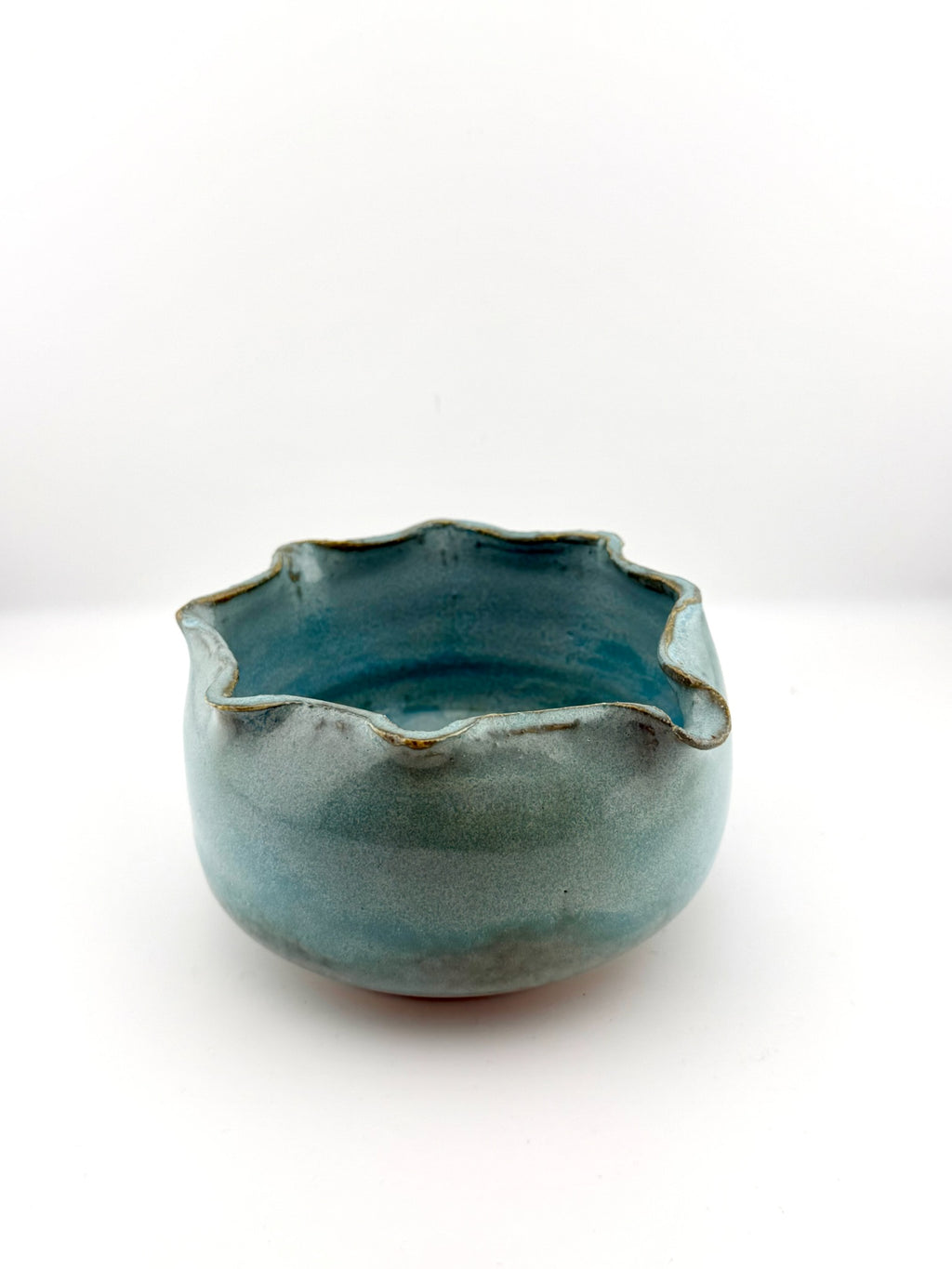 A handmade ceramic bowl with a scalloped, wavy rim, featuring a glossy teal blue-green glaze with subtle brown speckles around the edge, set against a plain white background.