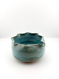 A handmade ceramic bowl with a scalloped, wavy rim, featuring a glossy teal blue-green glaze with subtle brown speckles around the edge, set against a plain white background.