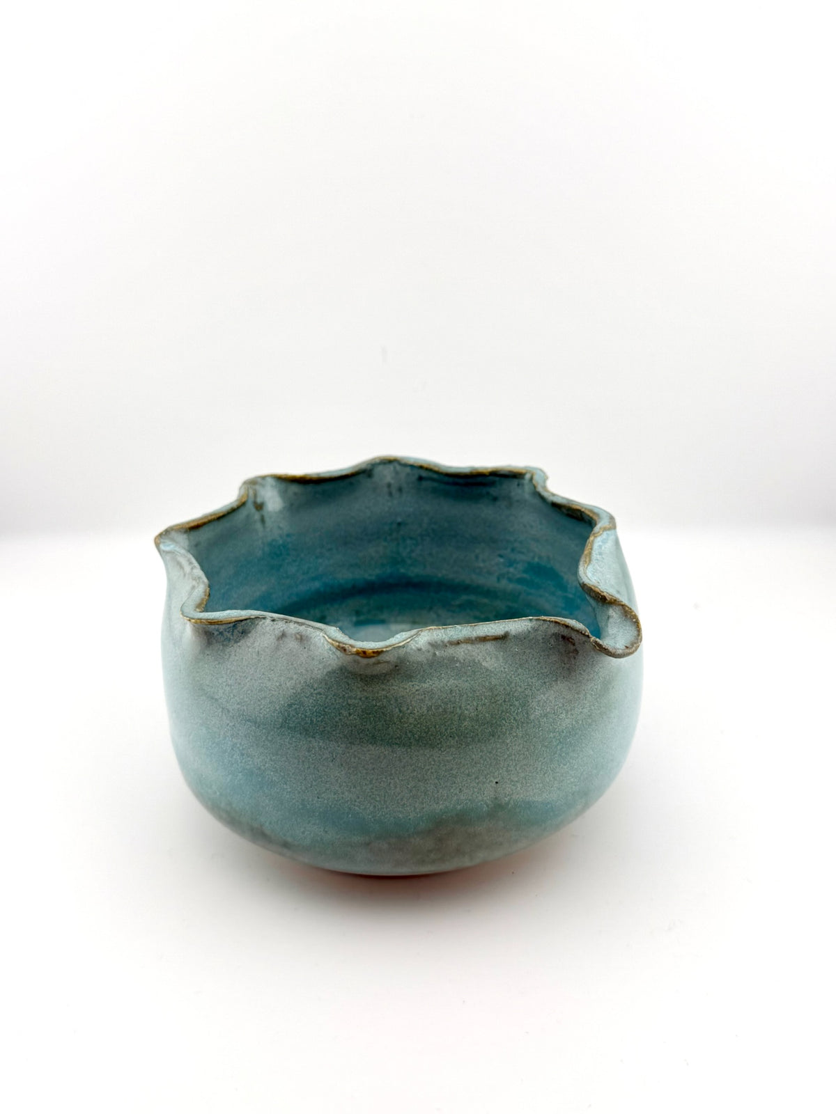 A handmade ceramic bowl with a scalloped, wavy rim, featuring a glossy teal blue-green glaze with subtle brown speckles around the edge, set against a plain white background.