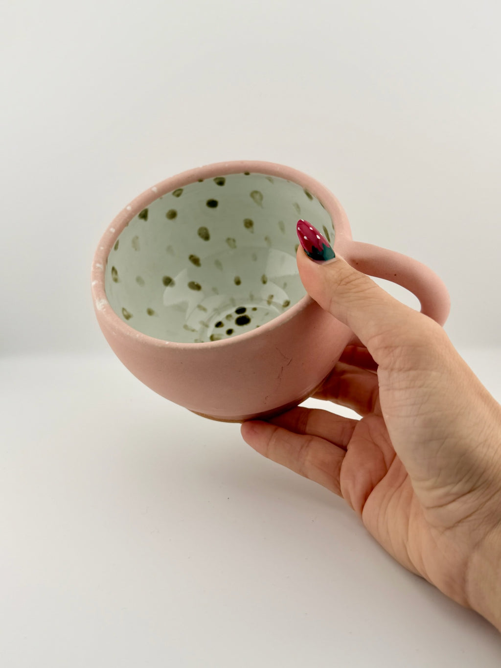 A hand holds a handmade pink ceramic mug with a speckled light green interior against a plain white background.