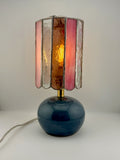 A vintage-style table lamp with a glossy dark blue glass base and a stained-glass shade featuring vertical panels of pink, amber-brown, and clear glass, illuminated from within.