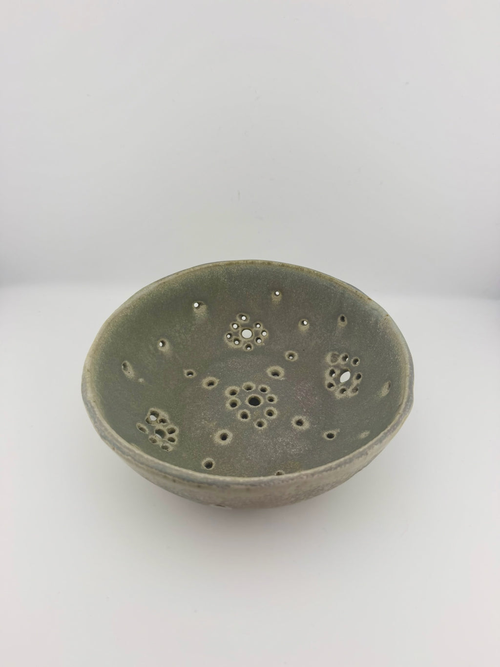 A handmade ceramic bowl with a textured, matte sage green glaze and decorative perforations on the inside, set against a plain white background.