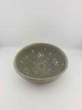A handmade ceramic bowl with a textured, matte sage green glaze and decorative perforations on the inside, set against a plain white background.